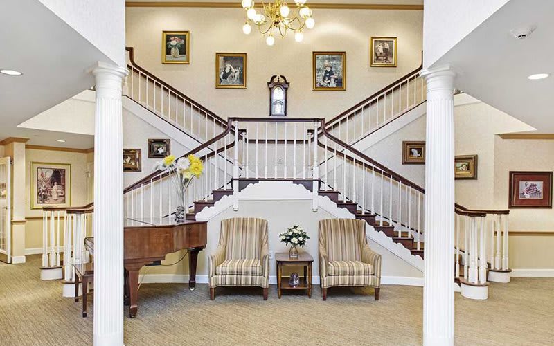 Elegant lobby with a grand staircase, adorned with framed pictures and a chandelier, featuring comfortable seating and a piano.
