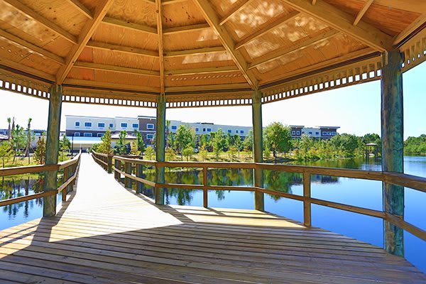 A wooden gazebo overlooks a tranquil lake with a surrounding bridge and modern buildings in the background.