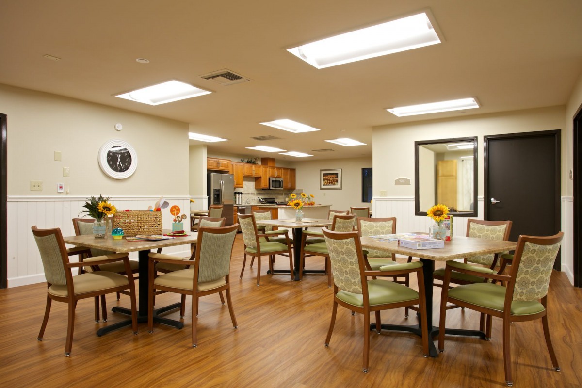 A well-lit dining room with multiple tables and chairs, featuring a small kitchen area in the background.
