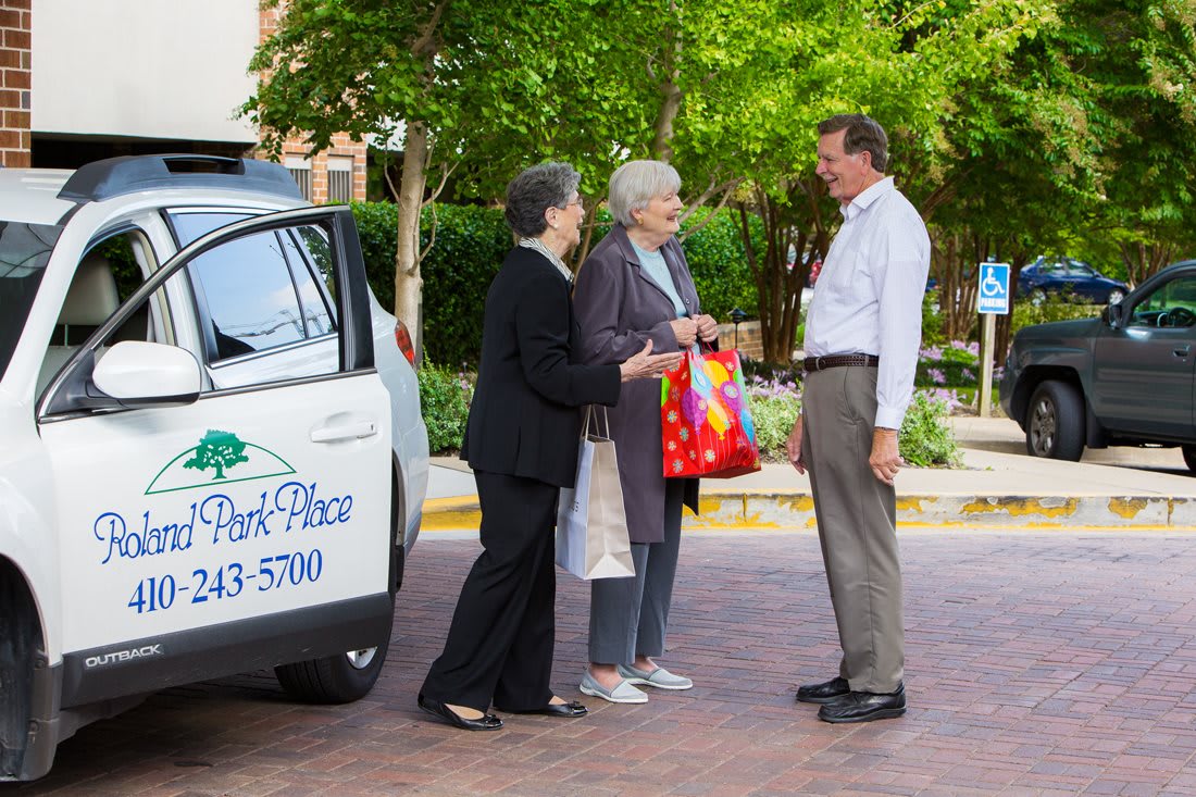 Two women talking with a man next to a car in a sunny outdoor setting.