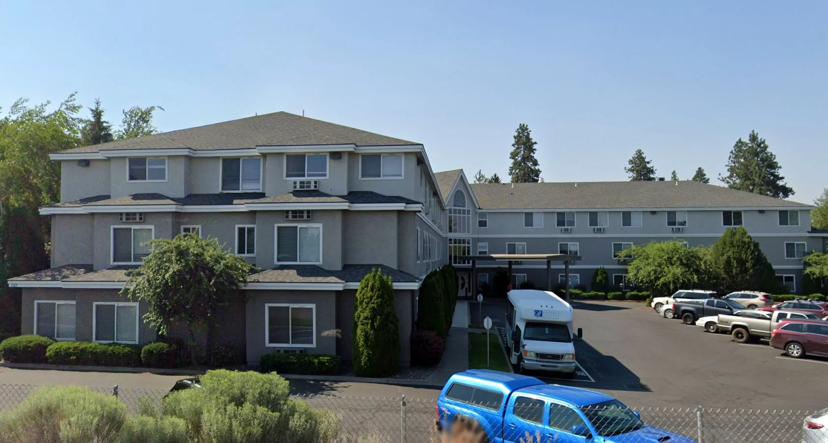 A residential building with a parking lot and several vehicles parked in front, set against a clear blue sky.