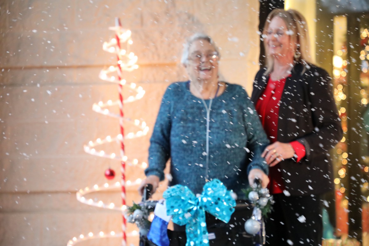An elderly woman with a walker, decorated for the holidays, is accompanied by a caregiver in a festive setting with lights and snow.