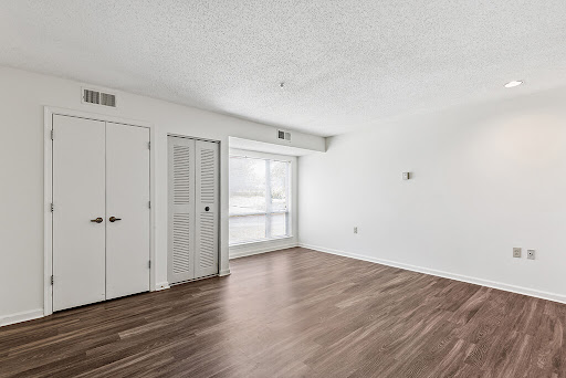 A spacious and empty interior room with white walls and wooden flooring, illuminated by natural light from the windows.