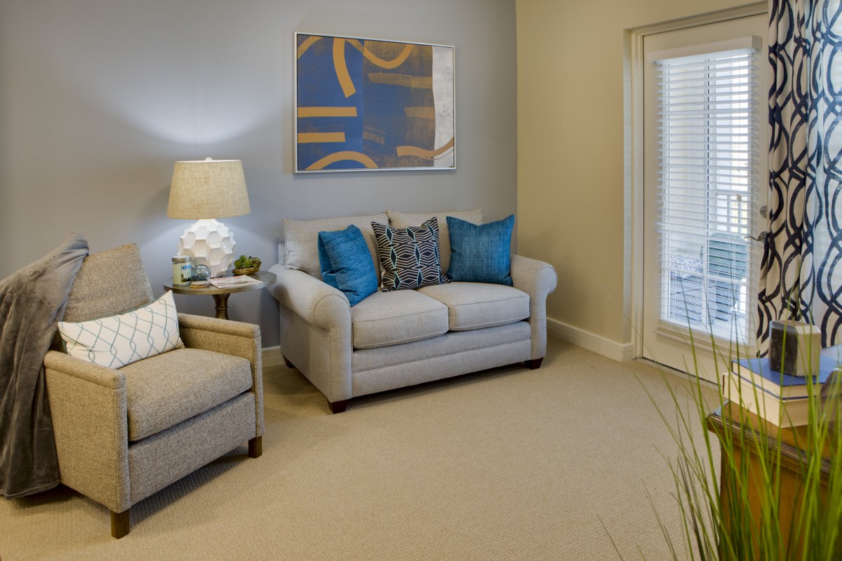 A cozy living room with a beige sofa and armchair, accented by blue and patterned pillows, features a floor lamp and abstract wall art.