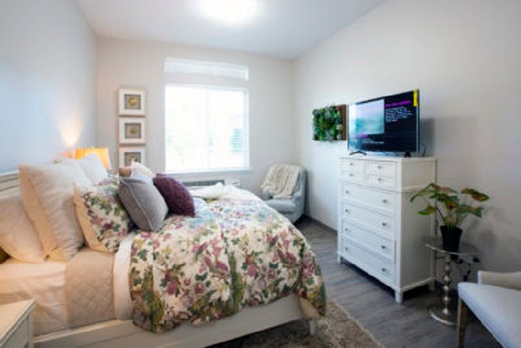 A cozy bedroom with floral bedding, a white dresser, and a television, featuring bright natural light from a window.