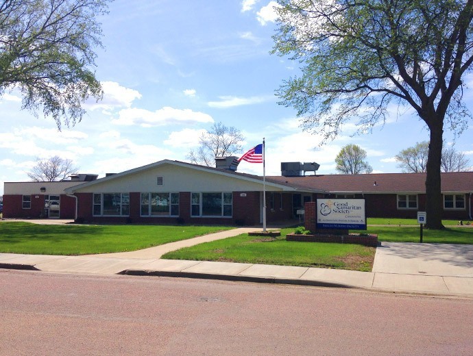 A single-story brick building with an American flag flying, surrounded by a green lawn and trees on a sunny day.