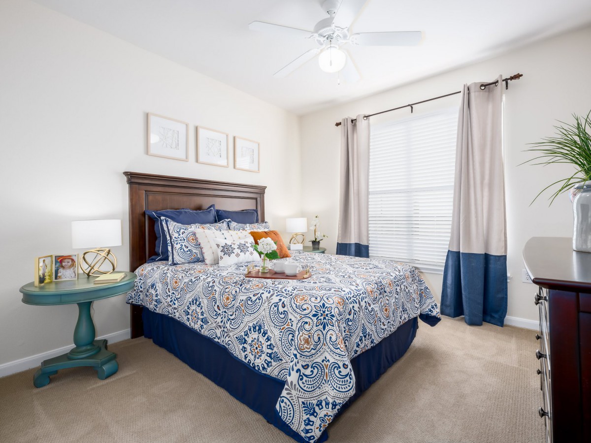 A well-decorated bedroom with a floral patterned bedspread, large window with curtains, and bedside lamps.