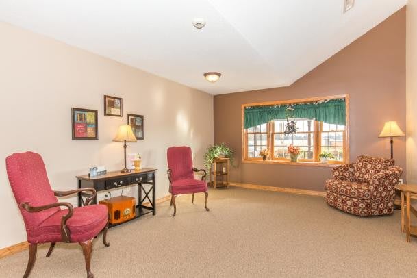 A cozy living room with armchairs, a desk with a lamp, and a large window allowing natural light.