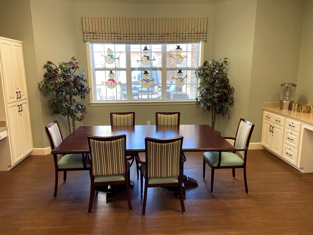 A cozy dining area featuring a wooden table surrounded by six chairs, with large windows and decorative plants on either side.