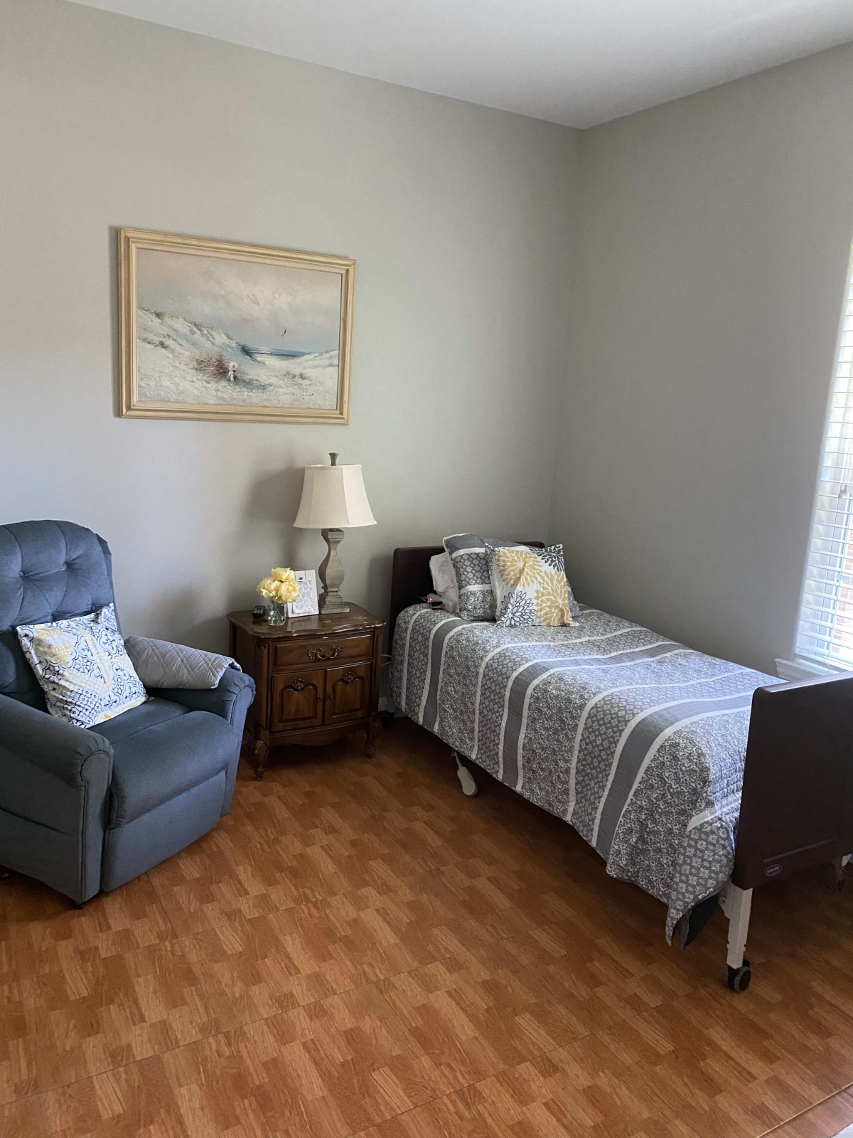 A cozy bedroom with a single bed, blue armchair, nightstand, lamp, and a painting on the wall.