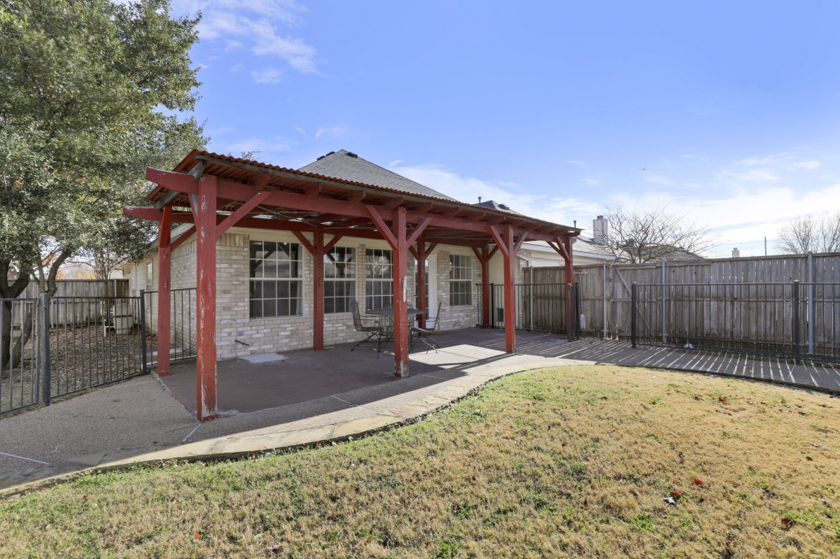 A backyard with a covered patio featuring a pergola, surrounded by a lawn and fenced area.