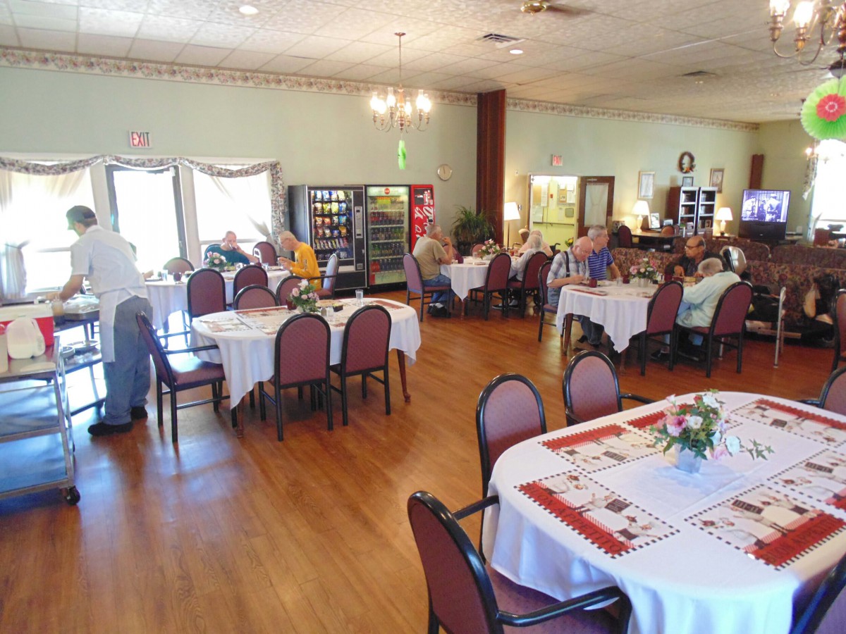 A casual dining area with several tables and chairs, occupied by people. Vending machines are visible in the background.