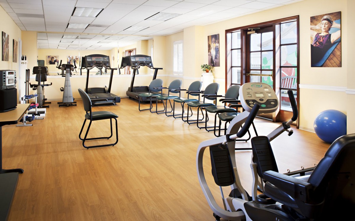 A fitness room with exercise equipment, including treadmills and chairs, on a wooden floor.