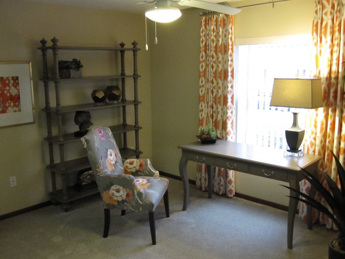 A cozy home office with a desk, upholstered chair, and shelving unit near a window with patterned curtains.