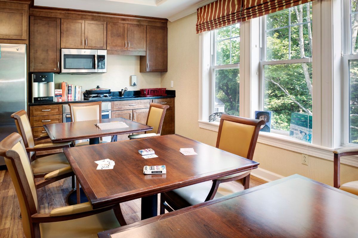 A cozy dining area with wooden tables and chairs, adjacent to a kitchen with wood cabinets and a microwave under natural light from large windows.