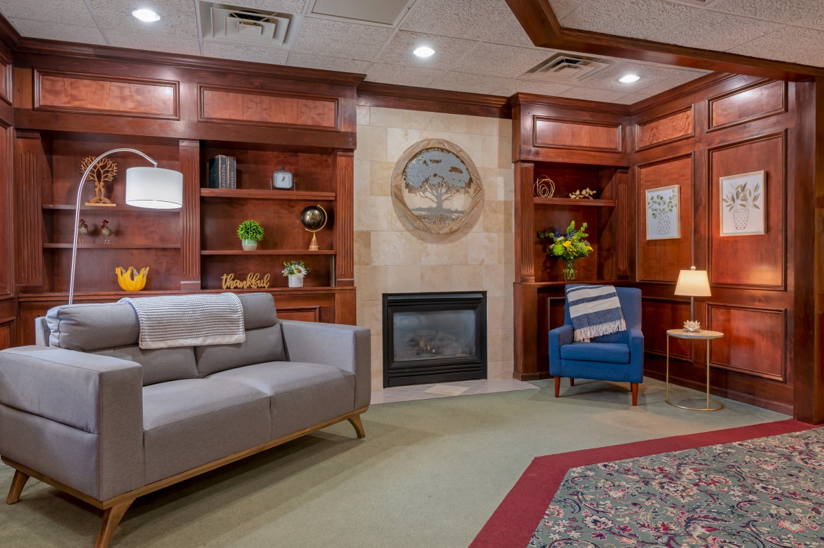A cozy living room with wood paneling, featuring a fireplace, sofa, armchair, and built-in bookshelves.