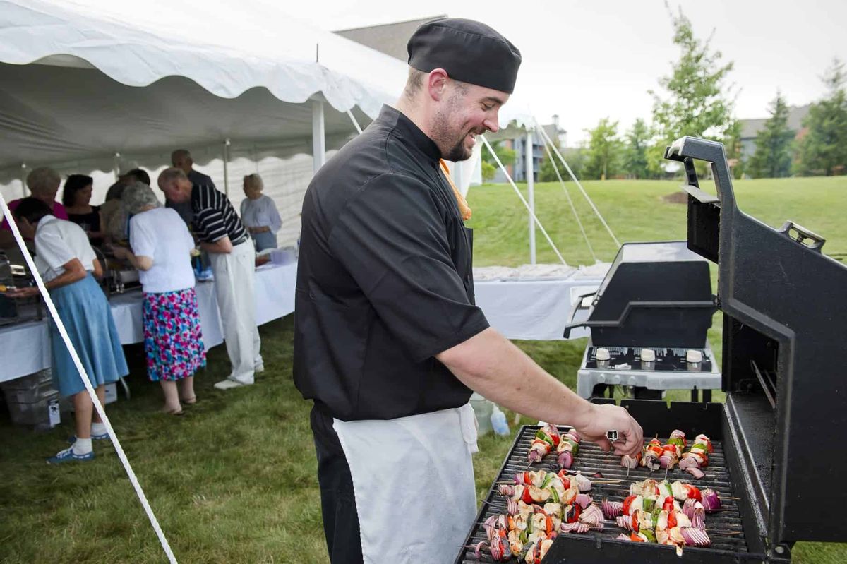A chef grills skewers at an outdoor event under a tent.