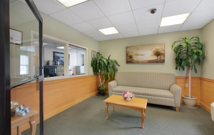 A cozy waiting room with a sofa, coffee table, and indoor plant, featuring a reception window and wall art.
