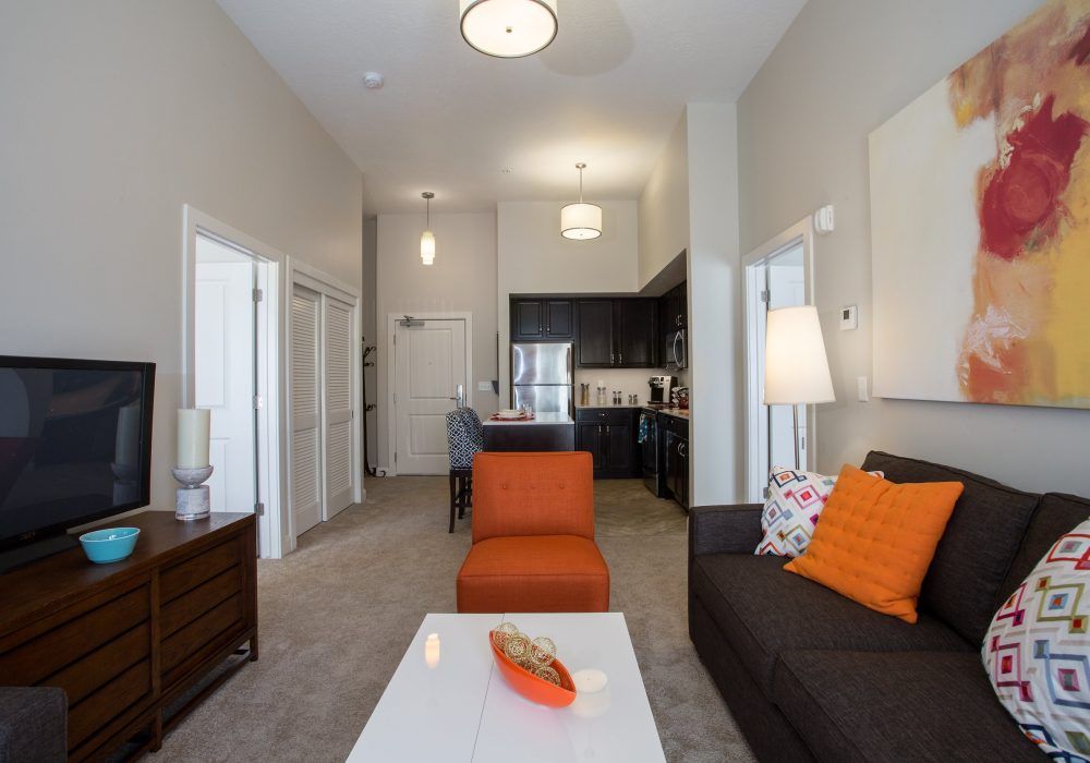 A modern living room with orange and dark furnishings, featuring a cozy seating area leading into an open kitchen.