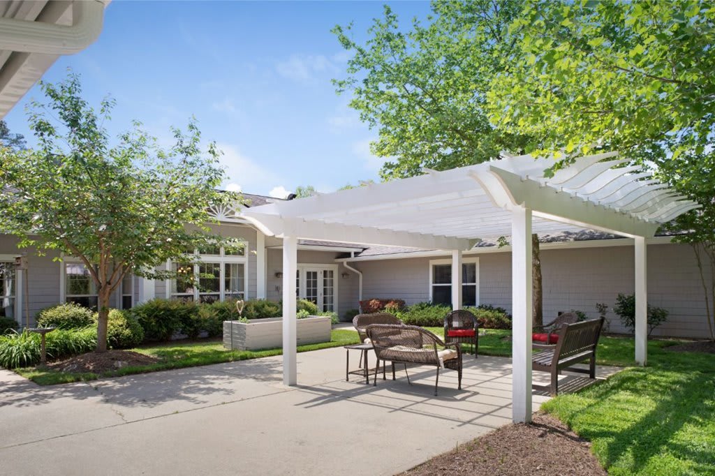 A sunny outdoor patio area with benches and a canopy, surrounded by trees and a building.