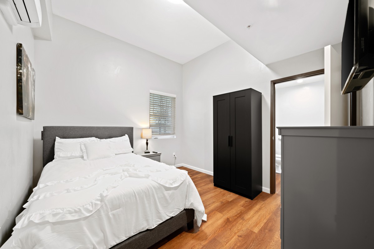 A minimalist bedroom with white walls and a wooden floor, featuring a bed and wardrobe.