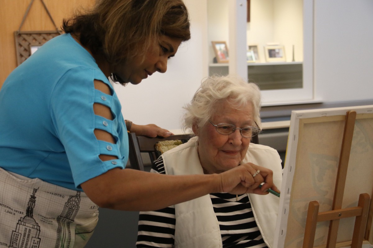 An elderly woman painting on a canvas with guidance from a teacher.