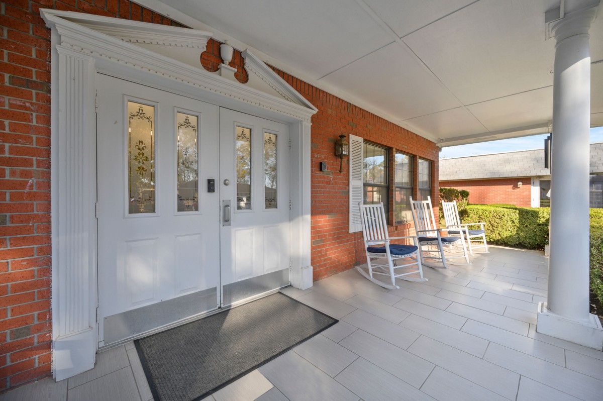 A welcoming porch with white double doors and rocking chairs against a brick wall.