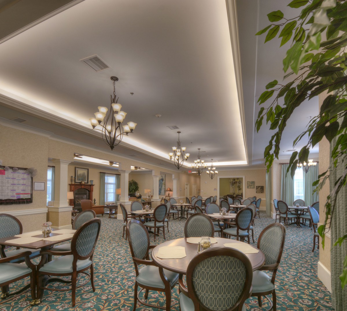 A well-decorated dining area with tables and chairs, illuminated by chandeliers, featuring carpeted flooring and greenery.