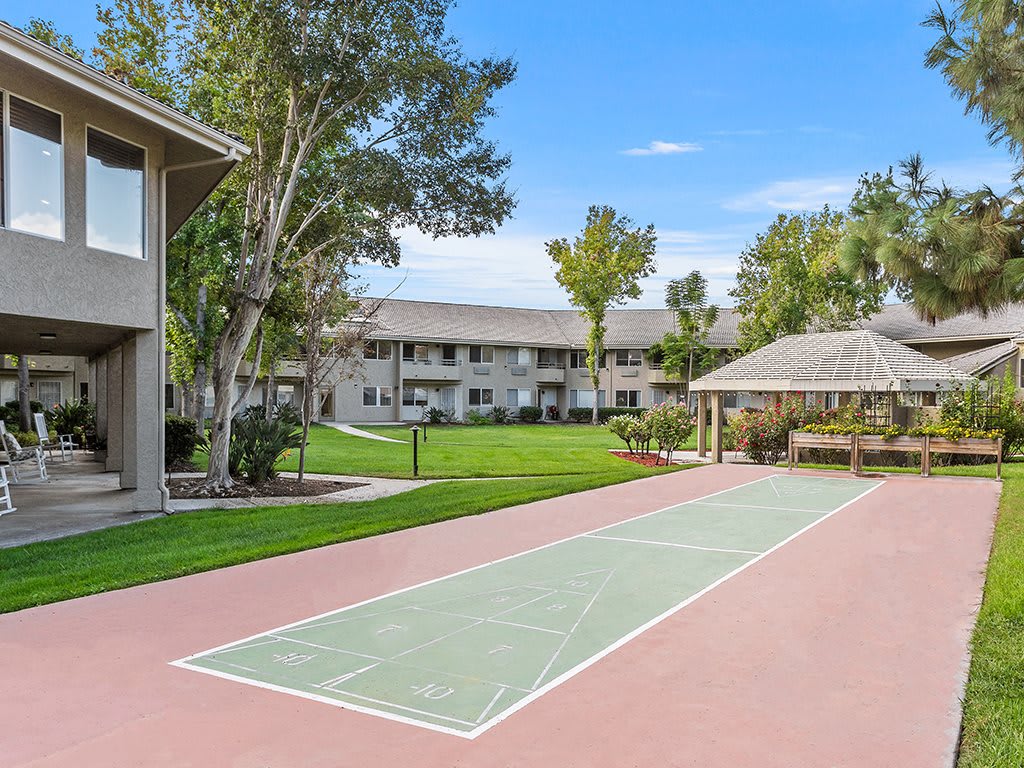 A well-maintained apartment complex featuring a shuffleboard court, surrounded by green lawns and trees.