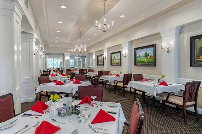 An elegant dining room with tables set with red napkins and chandeliers overhead.