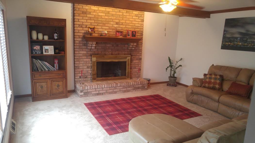 A cozy living room with a brick fireplace, a red plaid rug, and a ceiling fan.