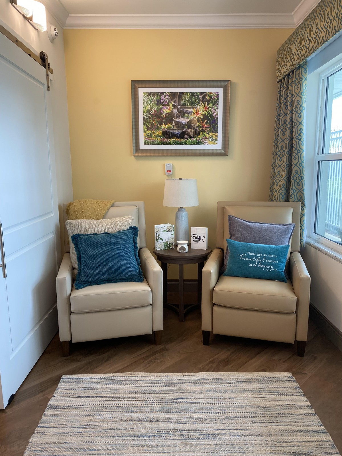 A cozy living room corner featuring two beige chairs with blue cushions, a small table with a lamp, and artwork on the wall by the window.