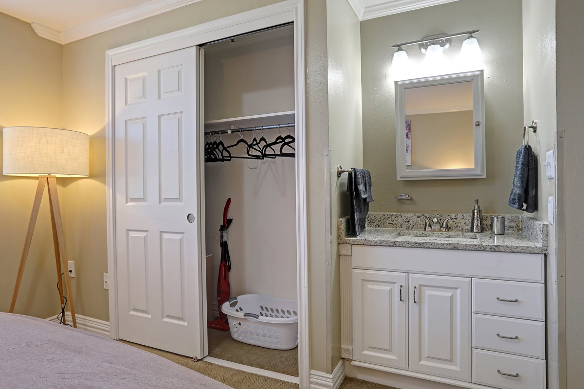 A bedroom with a small closet containing a vacuum cleaner and basket, adjacent to a vanity with a sink and mirror.