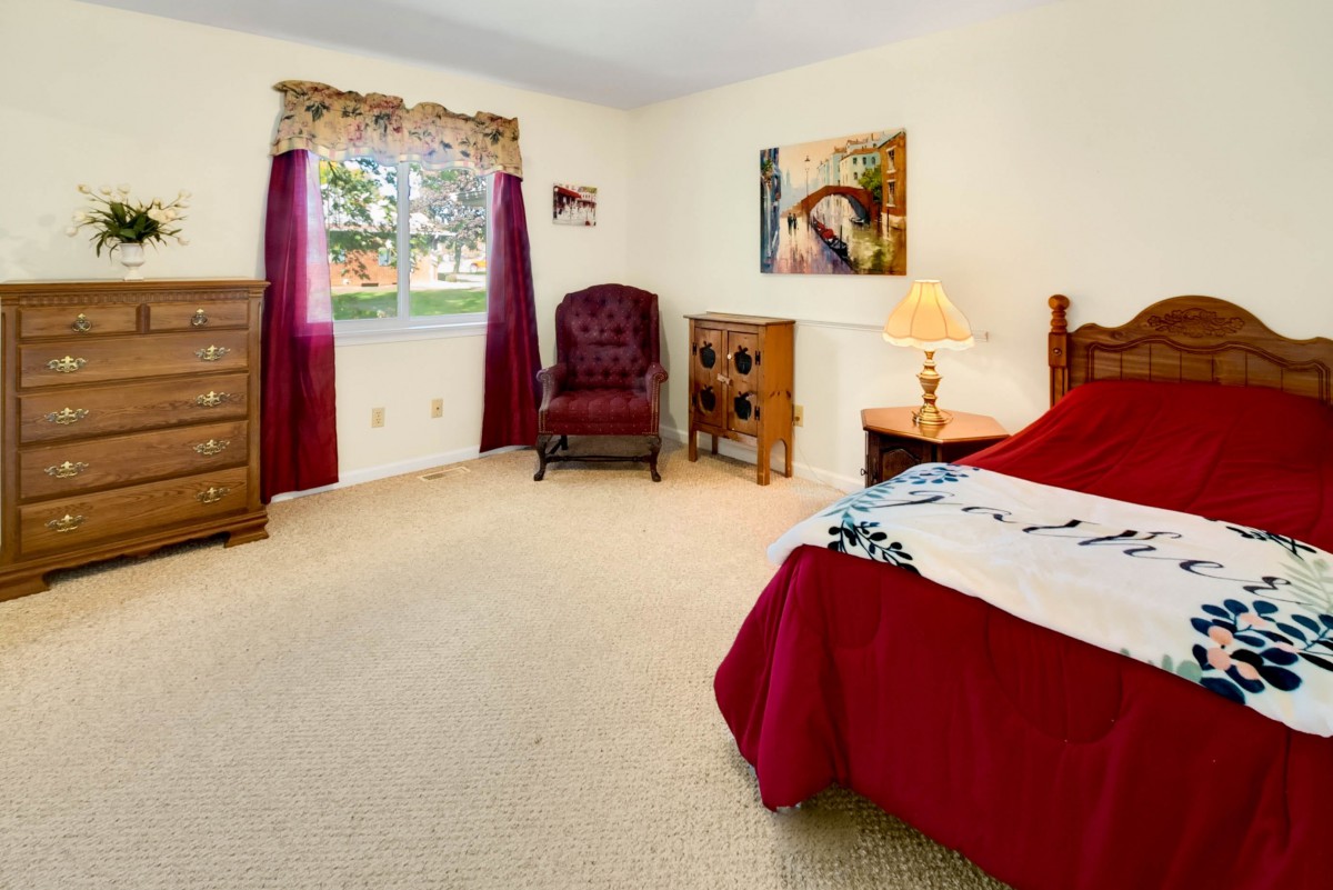 A cozy bedroom featuring a bed with a red comforter, a wooden dresser, and a window with curtains.