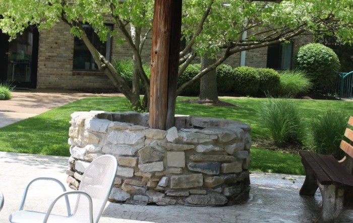 A stone well structure surrounds a tree in a garden with outdoor seating nearby.