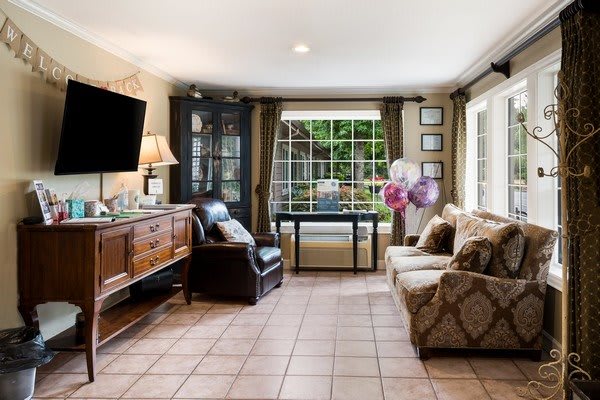 A cozy living room with tiled flooring, featuring a sofa, armchair, and a TV. Large windows allow natural light to illuminate the space.