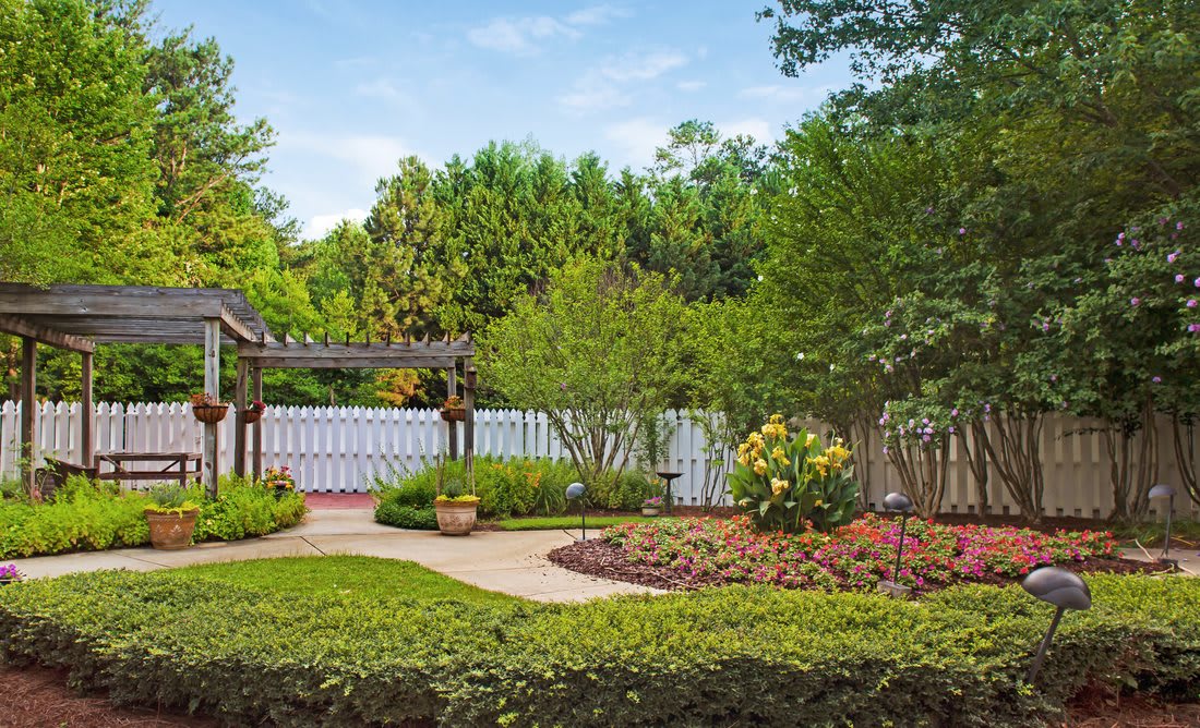 A beautifully landscaped garden with a paved pathway, surrounded by various plants and trees, enclosed by a white fence.