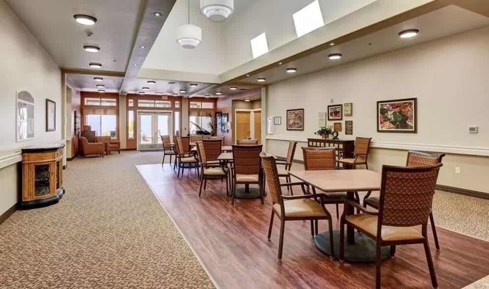 A spacious dining area with wooden flooring, tables, and chairs, illuminated by natural and ceiling lighting.