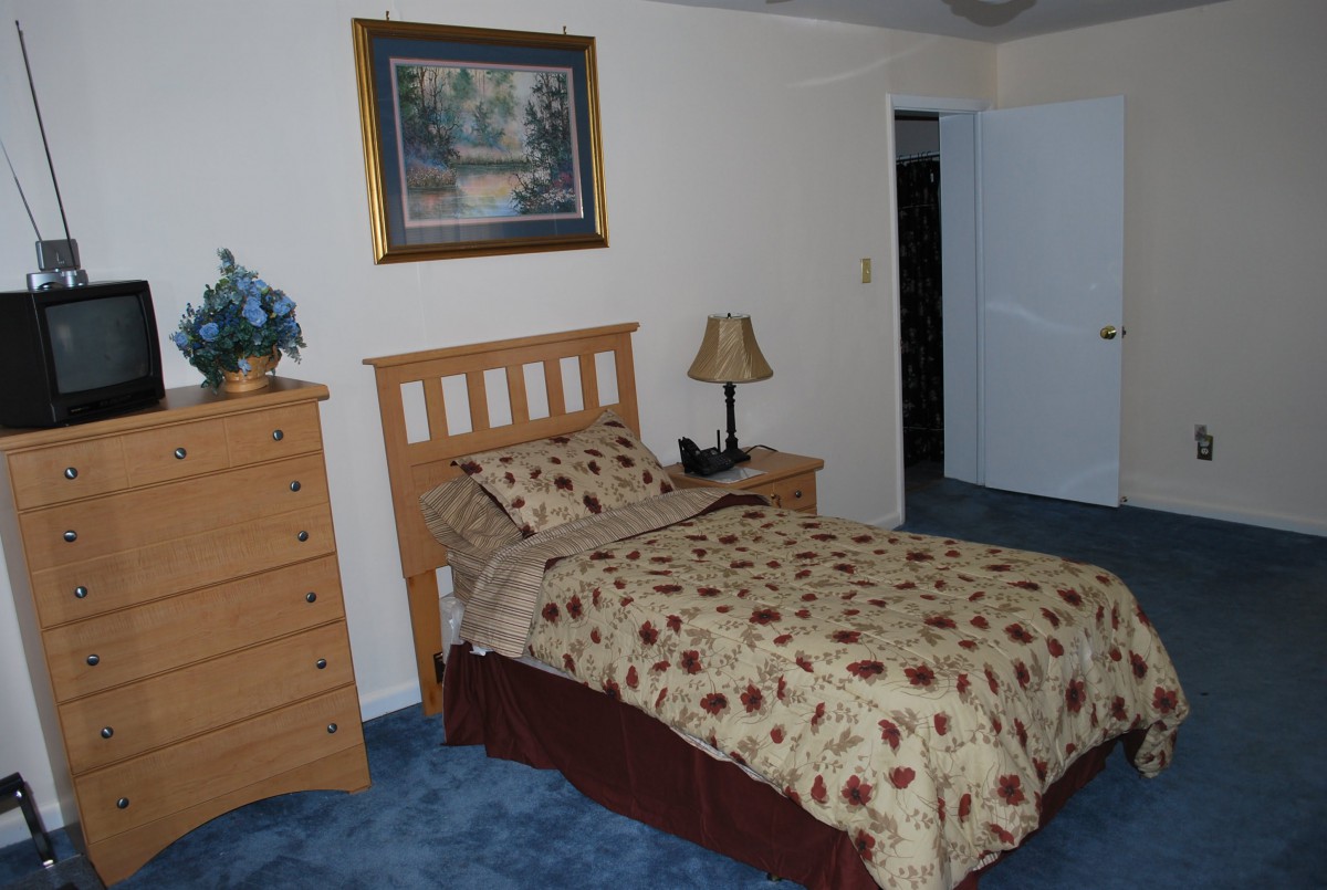 A neatly arranged bedroom with a single bed, dresser, lamp, and a painting on the wall.