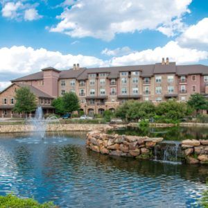 A scenic view of a building by a lake with fountains and a cloudy sky.