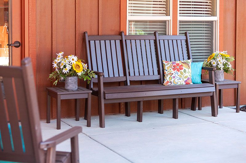 A cozy porch setting with a wooden bench, colorful decorative pillows, and a potted plant with flowers.
