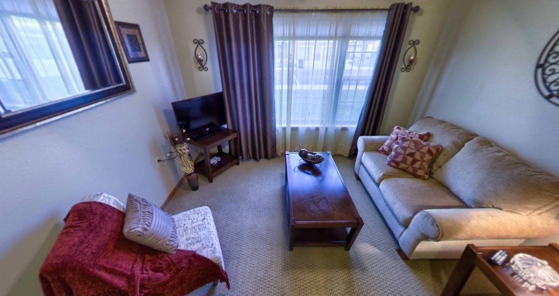 A cozy living room featuring a sofa, armchair, and coffee table, with a television in the corner near the window dressed with curtains.