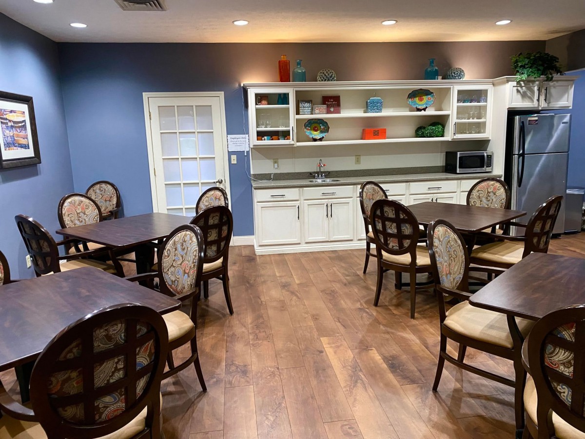 A cozy dining area with wooden tables and chairs, accompanied by a kitchenette and shelving against the wall.
