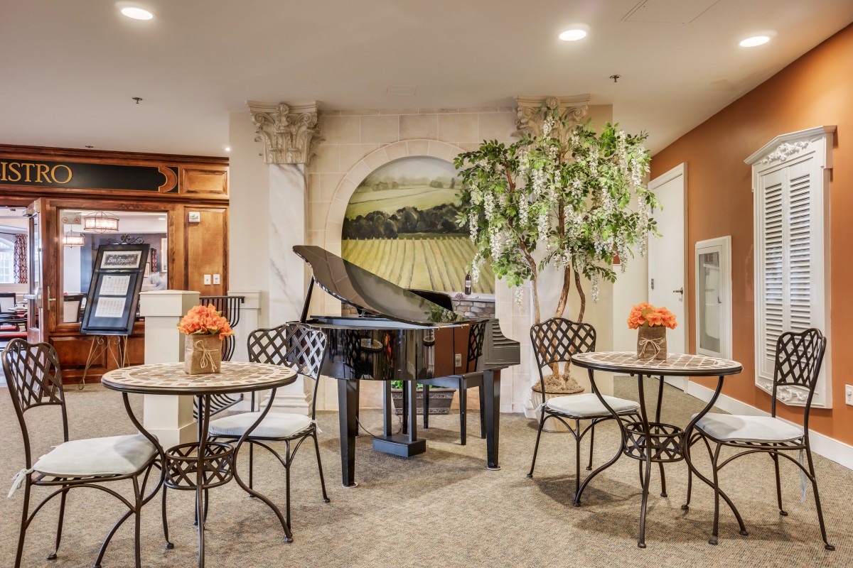 A cozy indoor setting featuring a grand piano surrounded by small tables and chairs, with decorative plants and artwork adorning the space.