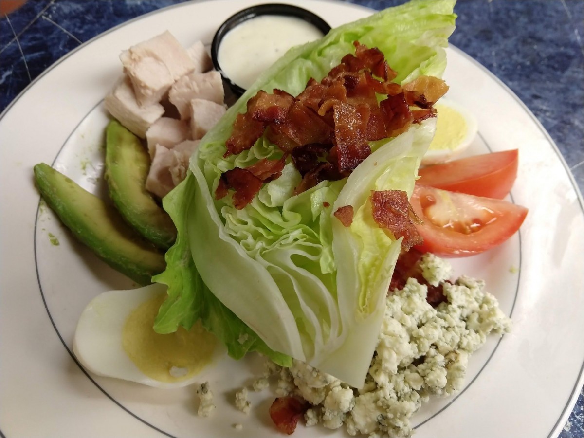 A wedge salad featuring bacon, avocado, tomato, chicken, blue cheese, lettuce, and egg.