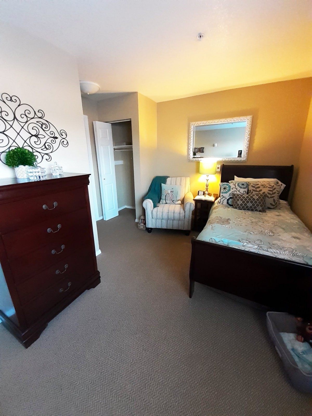 A cozy bedroom featuring a single bed, dresser, armchair, mirror, and lamp in warm lighting.