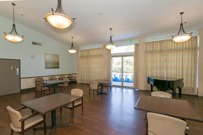 A dining area with tables, chairs, and a piano, featuring wooden floors and large windows with curtains.
