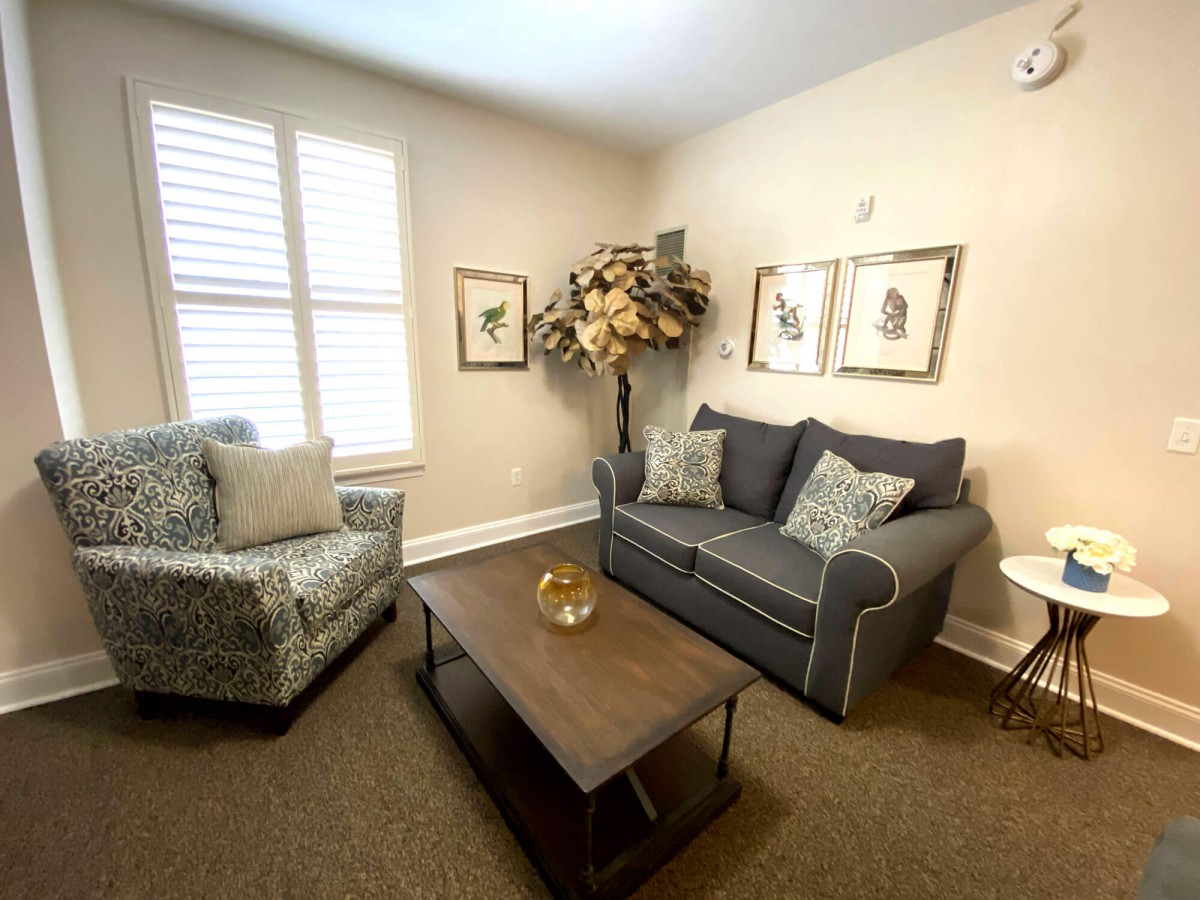 A cozy living room with a sofa, armchair, and coffee table, decorated with a potted plant and framed art on the walls.