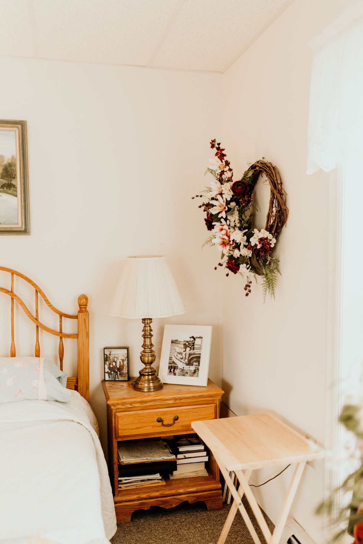 A cozy bedroom corner featuring a wooden nightstand with a lamp and photo frames, adorned with a floral wreath on the wall.