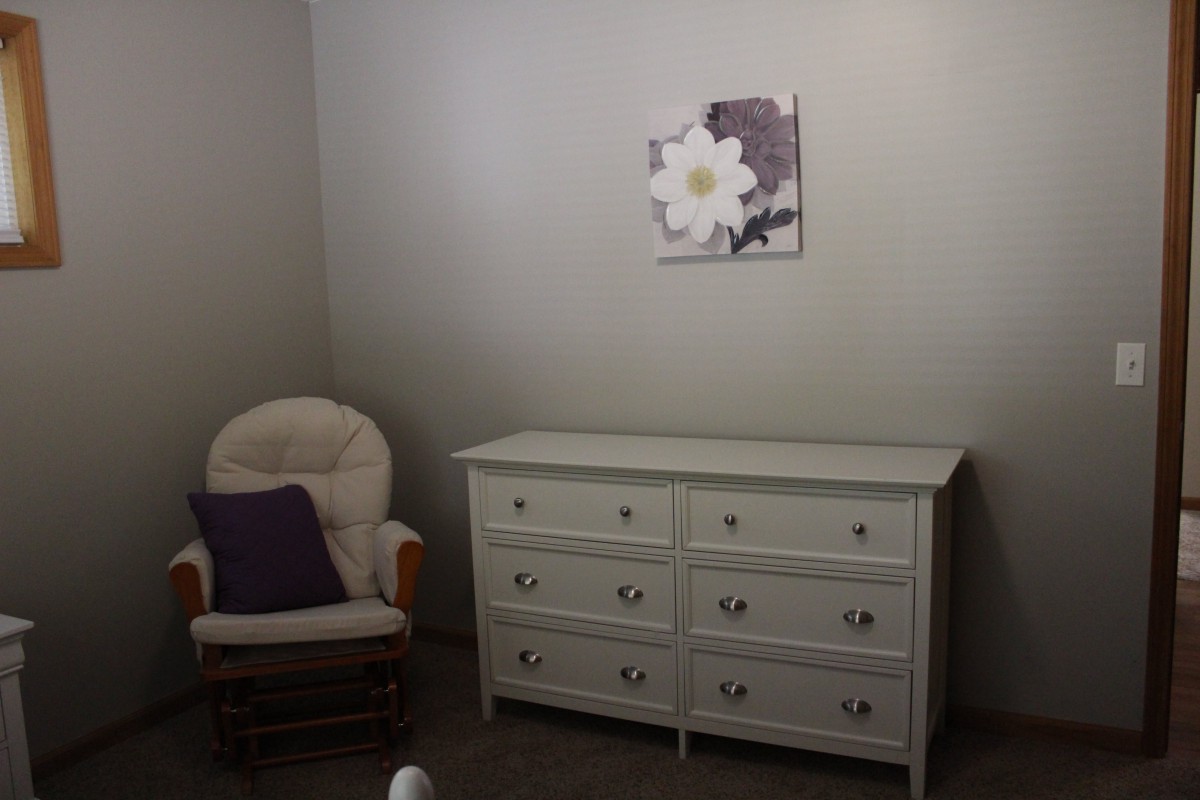 A minimalistic room featuring a white dresser, an armchair with a purple pillow, and a floral painting on the wall.